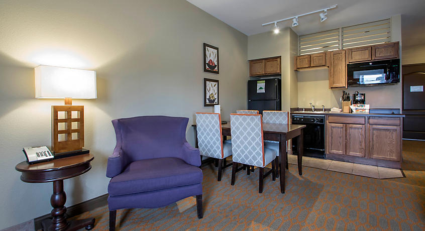 A cozy living space features a purple armchair and a wooden side table with a lamp. A dining table with four patterned chairs is positioned near a kitchenette equipped with dark appliances and cabinetry. Soft lighting enhances the inviting atmosphere of the room.