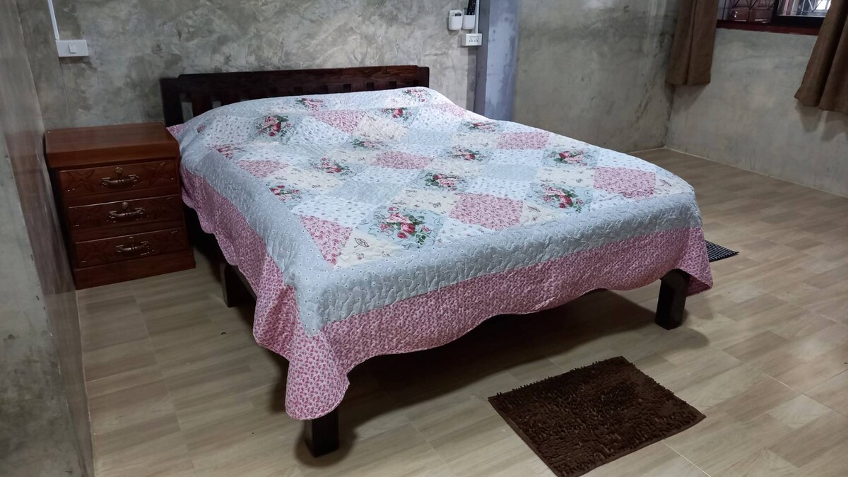 A spacious room is furnished with a large bed, covered in a patterned quilt featuring floral designs. Wooden side tables are located beside the bed, and the floor is covered in light-colored tiles, creating an inviting and tidy atmosphere.