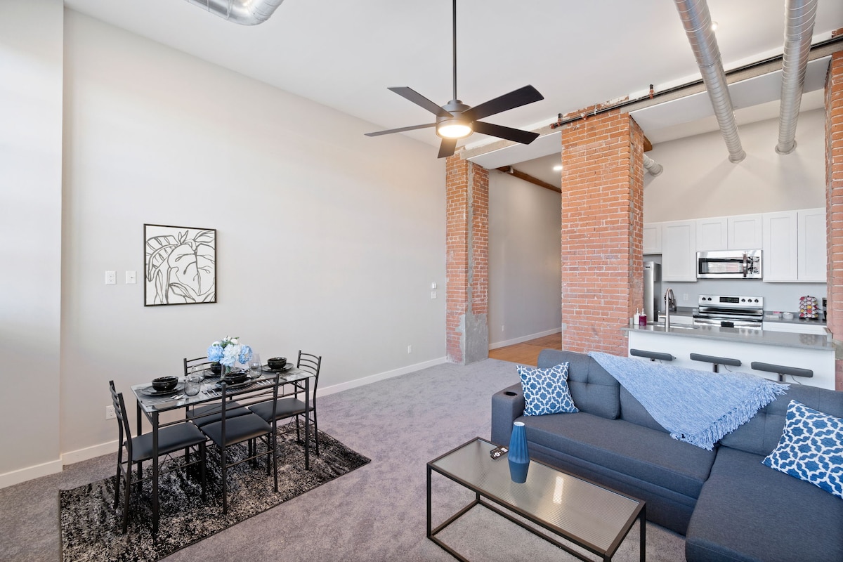 An open living area is highlighted by a modern ceiling fan and exposed brick walls. A dining table for six sits beside the comfortable sofa, adorned with decorative pillows. A fully equipped kitchen area is visible in the background, providing a casual and inviting space.
