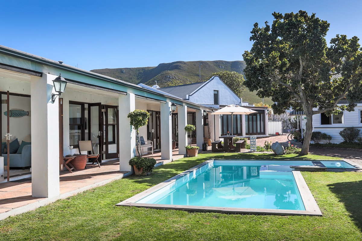 A serene outdoor area features a sparkling pool surrounded by neatly trimmed grass. Spacious lounge areas are positioned under covered patios, showcasing seated spaces with potted plants. A gentle slope of mountains rises in the background, under a clear blue sky.