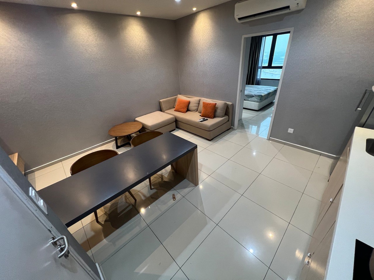 An open living area features a modern sofa with orange accent pillows, positioned beside two round tables. A sleek dining table is visible, along with large windows that allow natural light to enter. The bedroom area is partially visible in the background.
