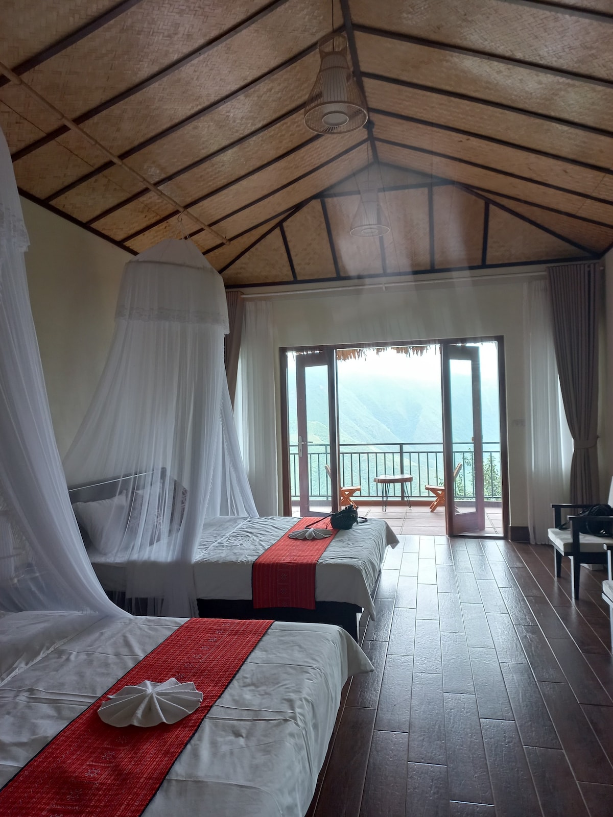 The room features two well-arranged beds draped with mosquito nets, creating a serene atmosphere. Large glass doors lead to a private balcony with seating, providing a view of the mountains and natural light. A warm wooden floor complements the airy ceiling design.