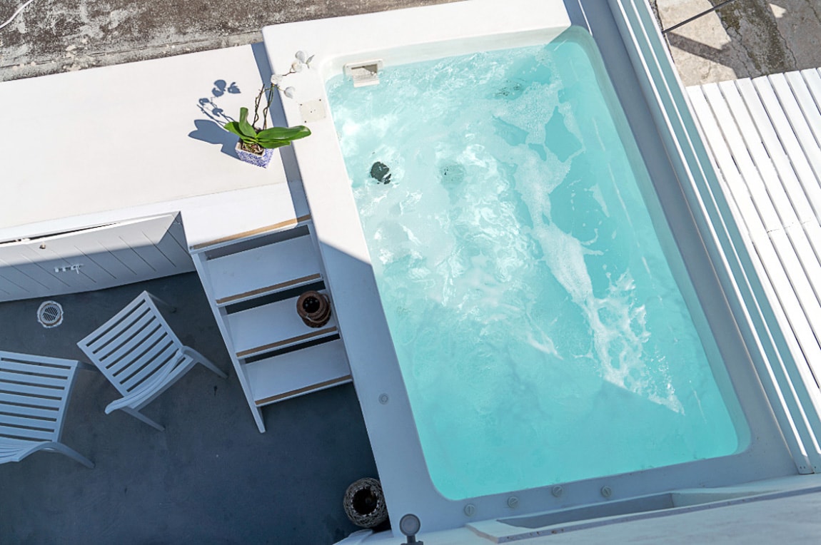 A private jacuzzi is surrounded by a clean and minimalistic patio space. Two white chairs are positioned nearby, enhancing the area. The tranquil water is visible as sunlight reflects off the surface, inviting relaxation.