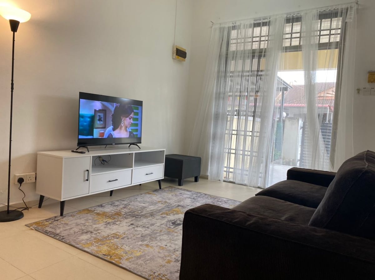 A cozy living area features a dark sofa facing a modern television mounted on a white media console. A soft area rug anchors the space, while natural light filters through large, sheer curtains that frame the windows, creating a relaxed atmosphere.