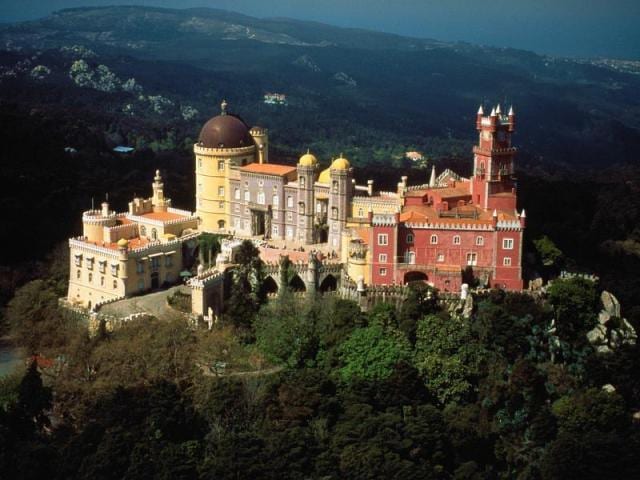 A historic castle complex is set within lush greenery, featuring distinct architectural styles and vibrant colors. Turrets and domes rise against the backdrop of rolling hills, creating an impressive silhouette that captures the essence of the region's heritage.