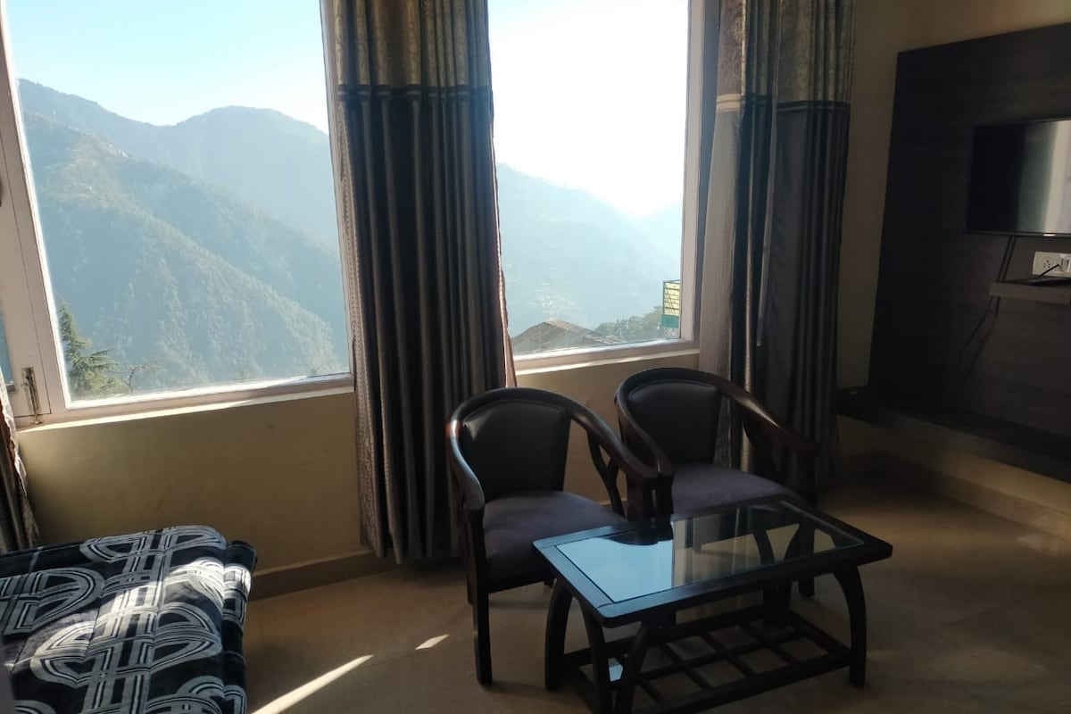 A bright living area features two comfortable chairs positioned around a glass coffee table. Large windows provide a view of distant mountains, with sunlight streaming in and illuminating the room. A mounted TV is visible on the wall, enhancing the relaxation space.