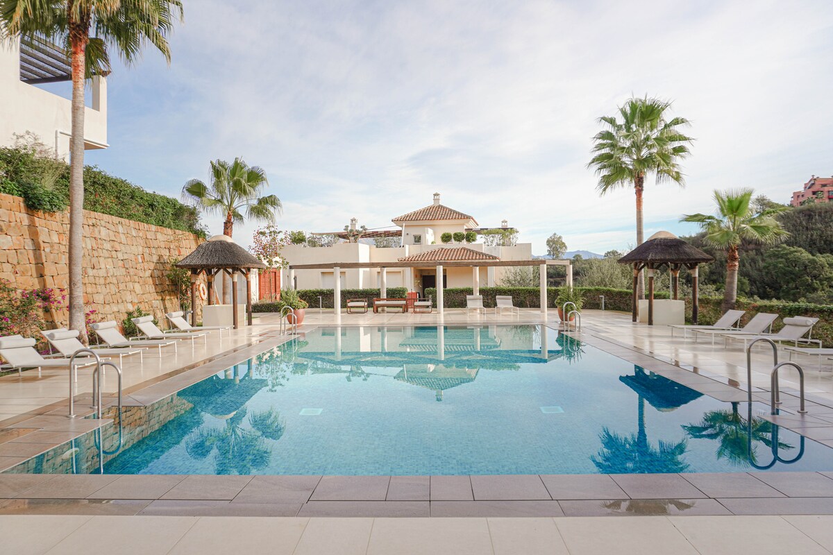 A spacious swimming pool is surrounded by lounge chairs, with shaded cabanas for relaxation. Lush palm trees and greenery frame the area, while a charming building with a tiled roof is visible in the background, contributing to the serene ambiance of the setting.