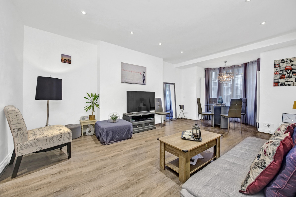 The living area features contemporary furnishings, including a spacious sofa and a wooden coffee table. A wall-mounted TV is positioned in front of the sofa, with a dining space visible in the background. Natural light fills the room through large windows, enhancing the open layout.