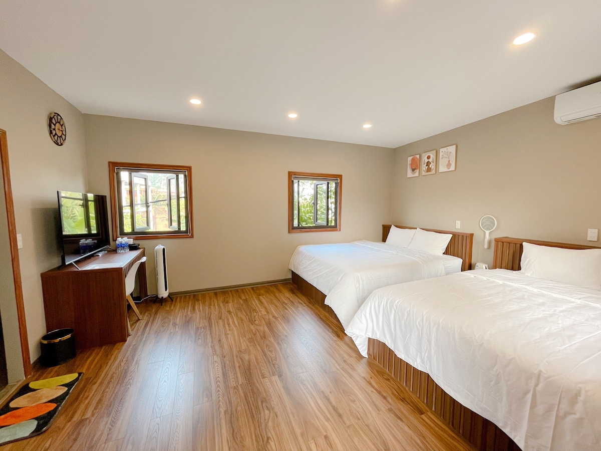 A spacious room is featured with two comfortable beds, positioned against a warm-toned wall. Natural light enters through two windows, enhancing the inviting atmosphere. A television is mounted on the wall, with a wooden table and decorative items nearby, creating a functional yet stylish design.