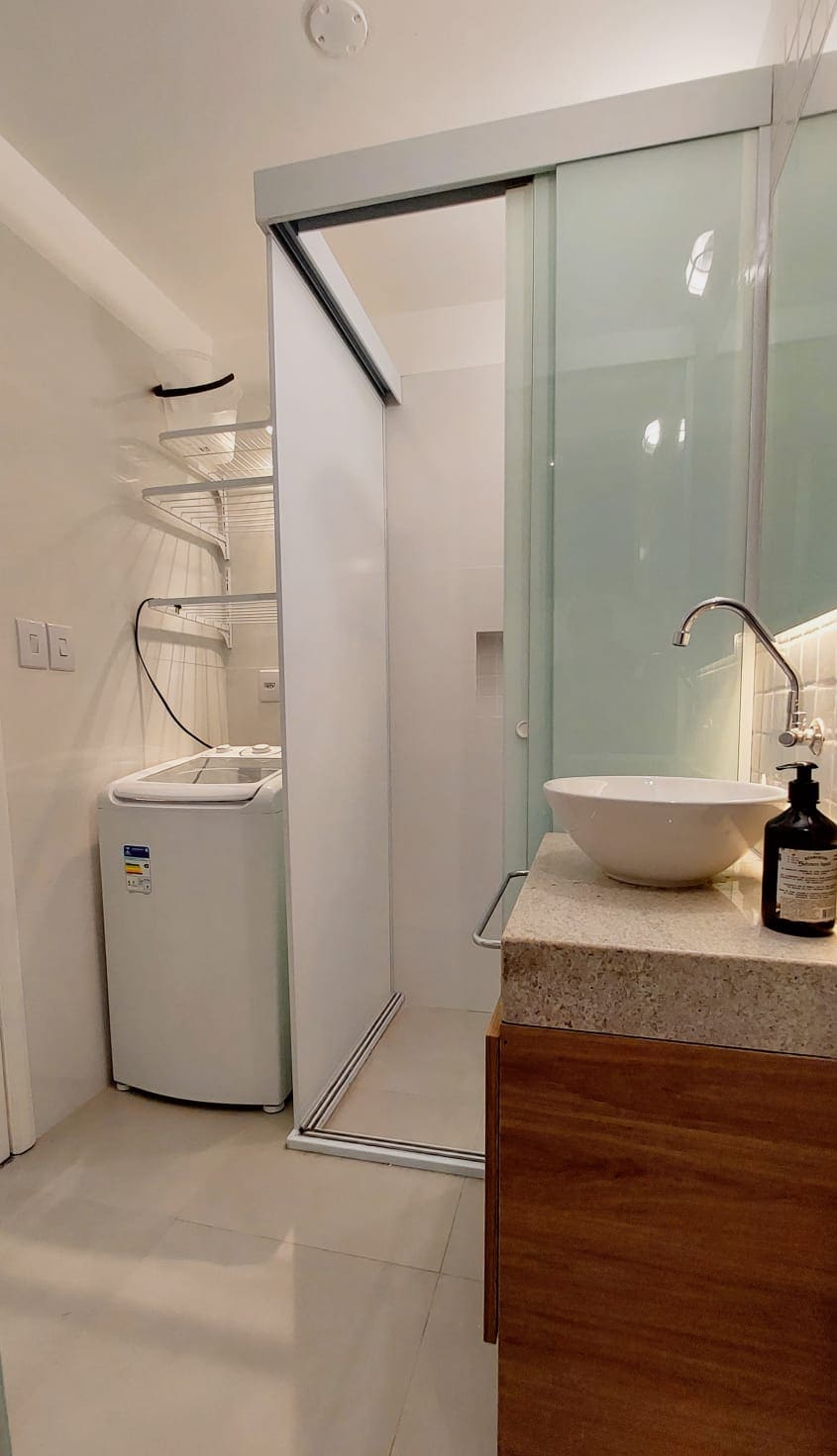 A modern bathroom is displayed, featuring a glass-enclosed shower area, a neatly arranged countertop with a round sink, and a washing machine present against the wall. Soft lighting highlights the clean lines and minimalist design of the space.