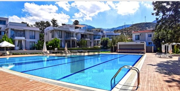 A large outdoor swimming pool is surrounded by sun loungers and umbrellas, set against a backdrop of modern villas and lush greenery. The clear water reflects the blue sky and distant mountains, providing a serene atmosphere for relaxation.