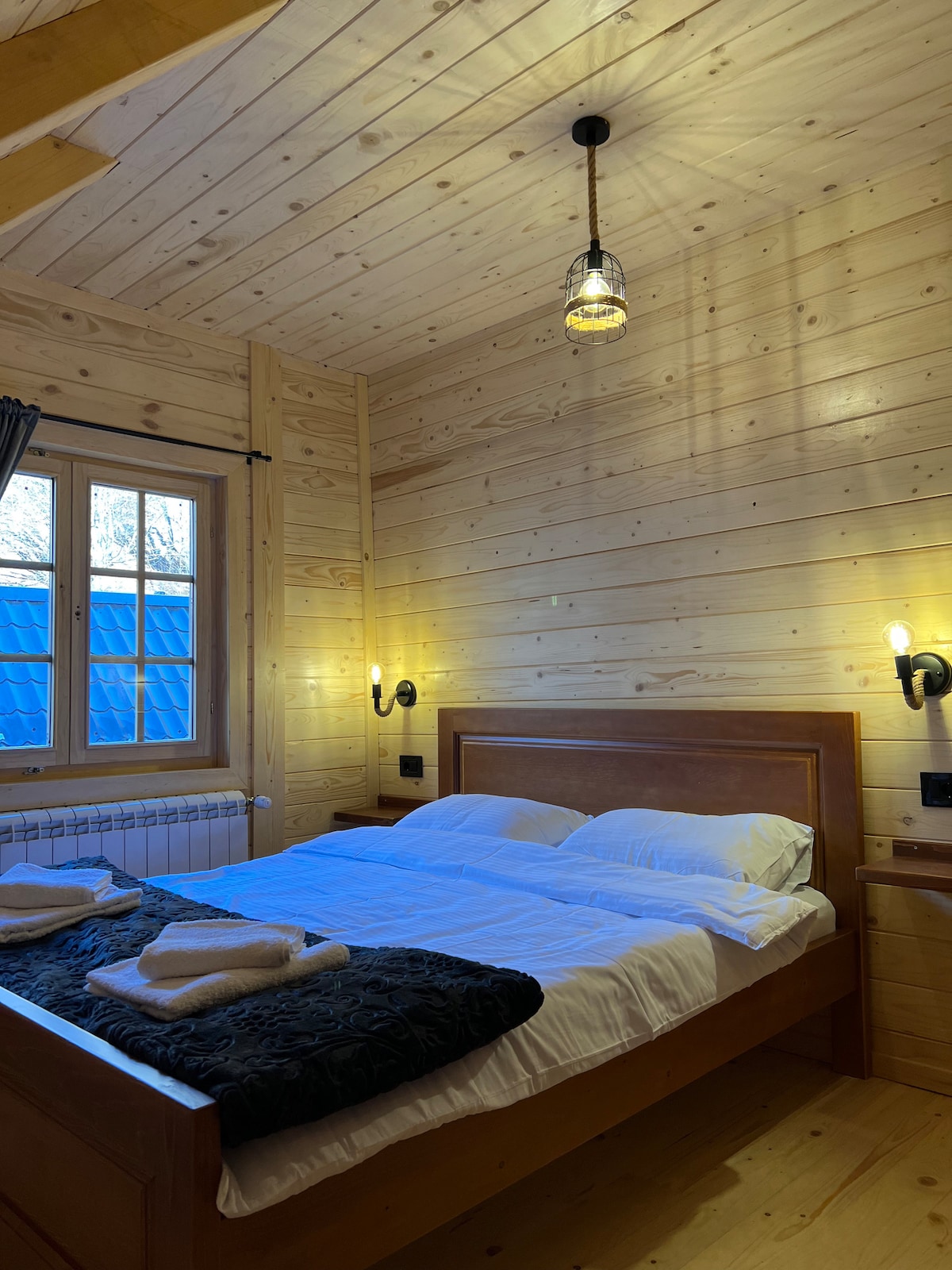 A warm wooden interior features a spacious bed adorned with neatly arranged linens and towels. Soft lighting emanates from wall sconces, enhancing the inviting atmosphere. A window allows natural light to enter, illuminating the room's natural wood accents.