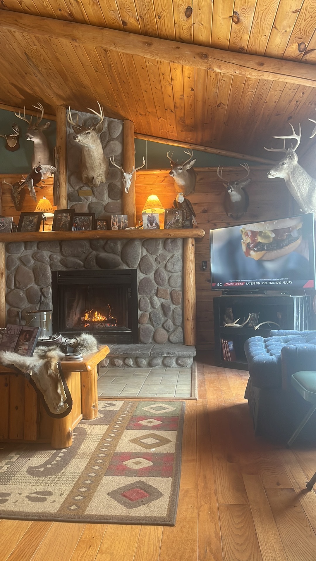 A spacious living area features a stone fireplace, providing warmth and a rustic charm. Taxidermy mounts decorate the walls, showcasing various wildlife. Comfortable seating is arranged around a wooden coffee table. A large television is positioned nearby, enhancing the entertainment options.