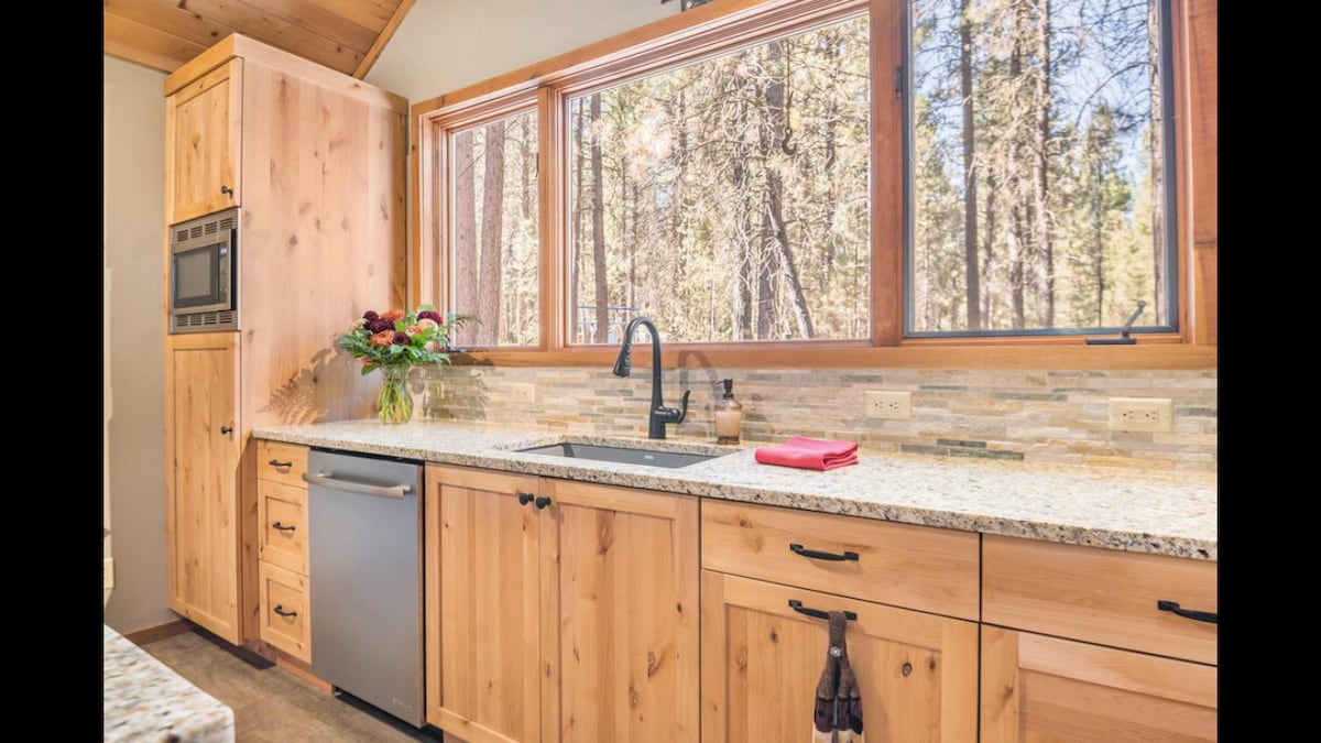 A well-appointed kitchen is featured, showcasing natural wood cabinetry and a granite countertop. Large windows provide views of the surrounding trees, allowing ample natural light. A decorative arrangement of flowers and a red cloth add subtle color to the space.