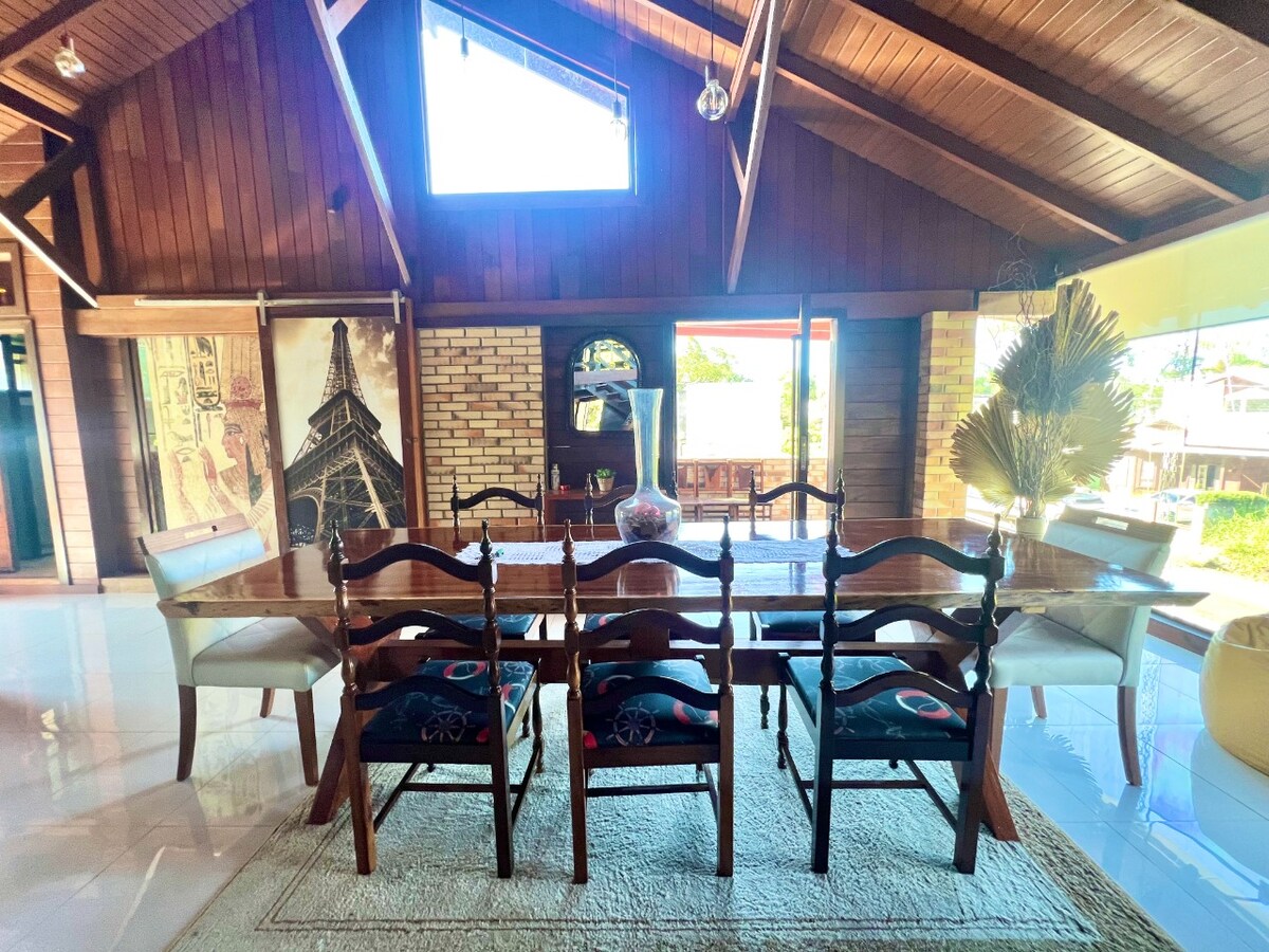 An open dining area features a large wooden table surrounded by six chairs. Natural light fills the space through a high window, highlighting the wooden beams of the vaulted ceiling. A plush rug is located underneath the table.