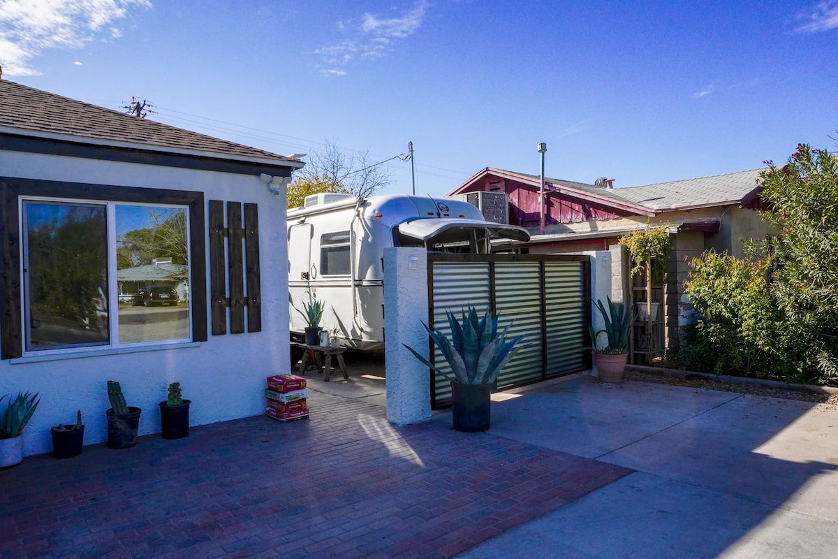 A vintage 1977 Avion is parked in a driveway, showcasing its metallic exterior against a clear blue sky. Surrounding plants add greenery, while the entrance is framed by a modern garage door and a white wall. Nearby structures reflect a blend of historic and contemporary architecture.