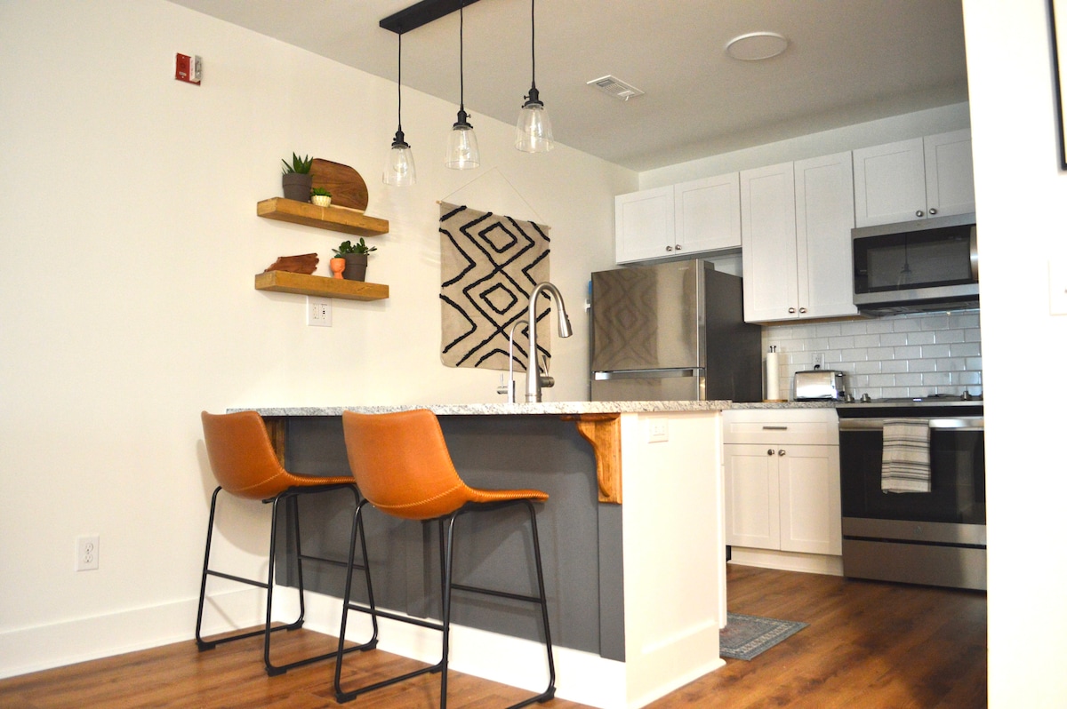 A modern kitchen area features a granite countertop with two stylish bar stools. Above, three pendant lights provide illumination. Open shelves are adorned with plants and decorative items. Stainless steel appliances and white cabinetry create a sleek and functional space.