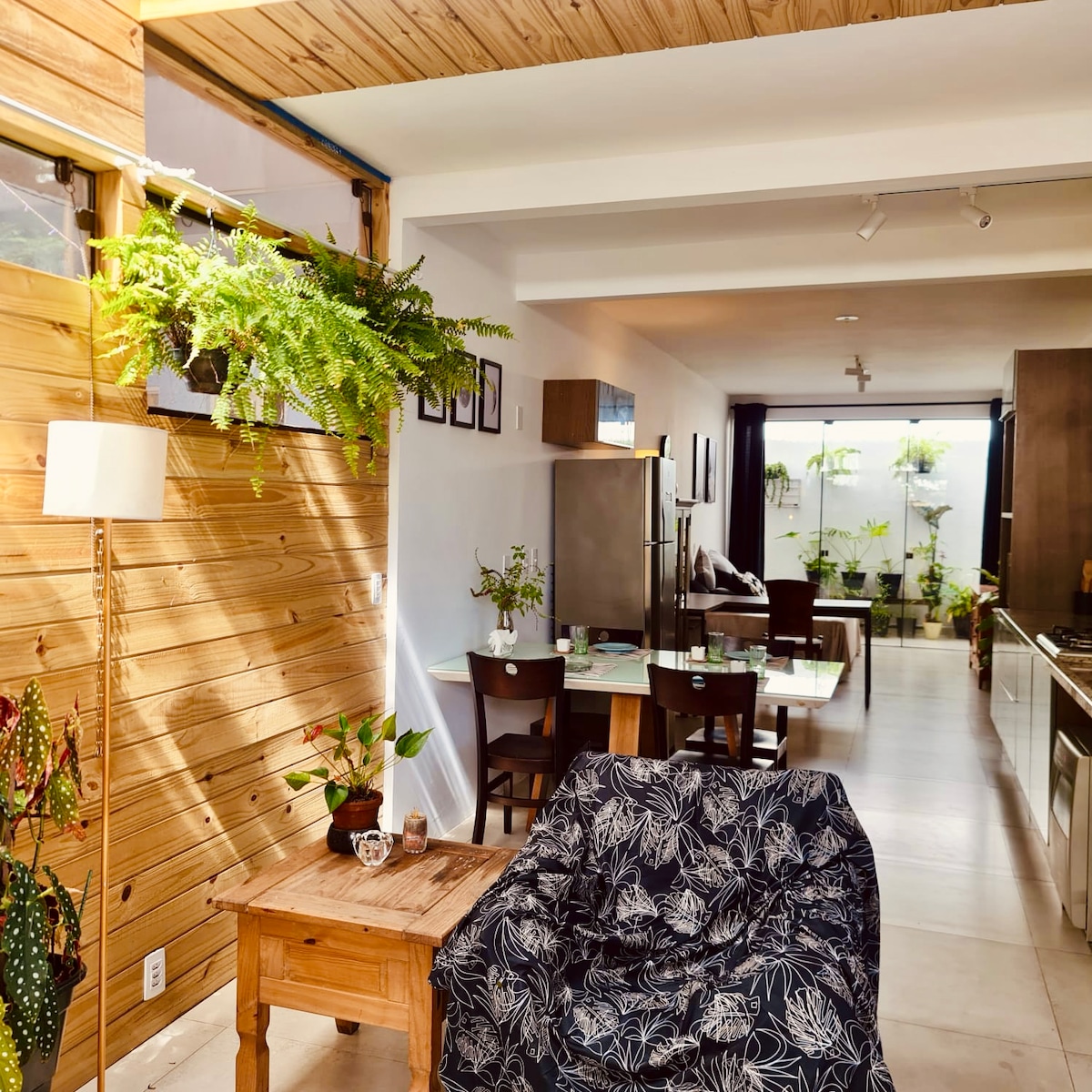 A well-lit living area features a comfortable seating arrangement with a patterned throw draped over the chair. The space incorporates natural elements with indoor plants and large windows, while the open layout connects the kitchen and dining areas, creating a seamless flow throughout.