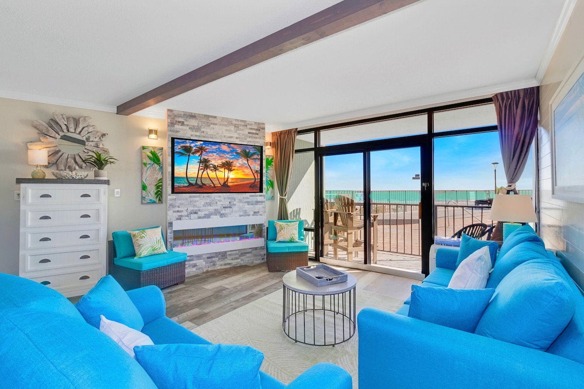 A spacious living area features bright blue seating arranged around a circular table. A flat-screen TV displays a sunset beach scene against a stone accent wall. Floor-to-ceiling glass doors open to an outdoor balcony, providing expansive views of the ocean.