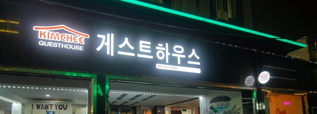 The guesthouse exterior is prominently displayed with a bright sign featuring both English and Korean text. Soft lighting accents the entrance, contributing to a welcoming appearance at night.