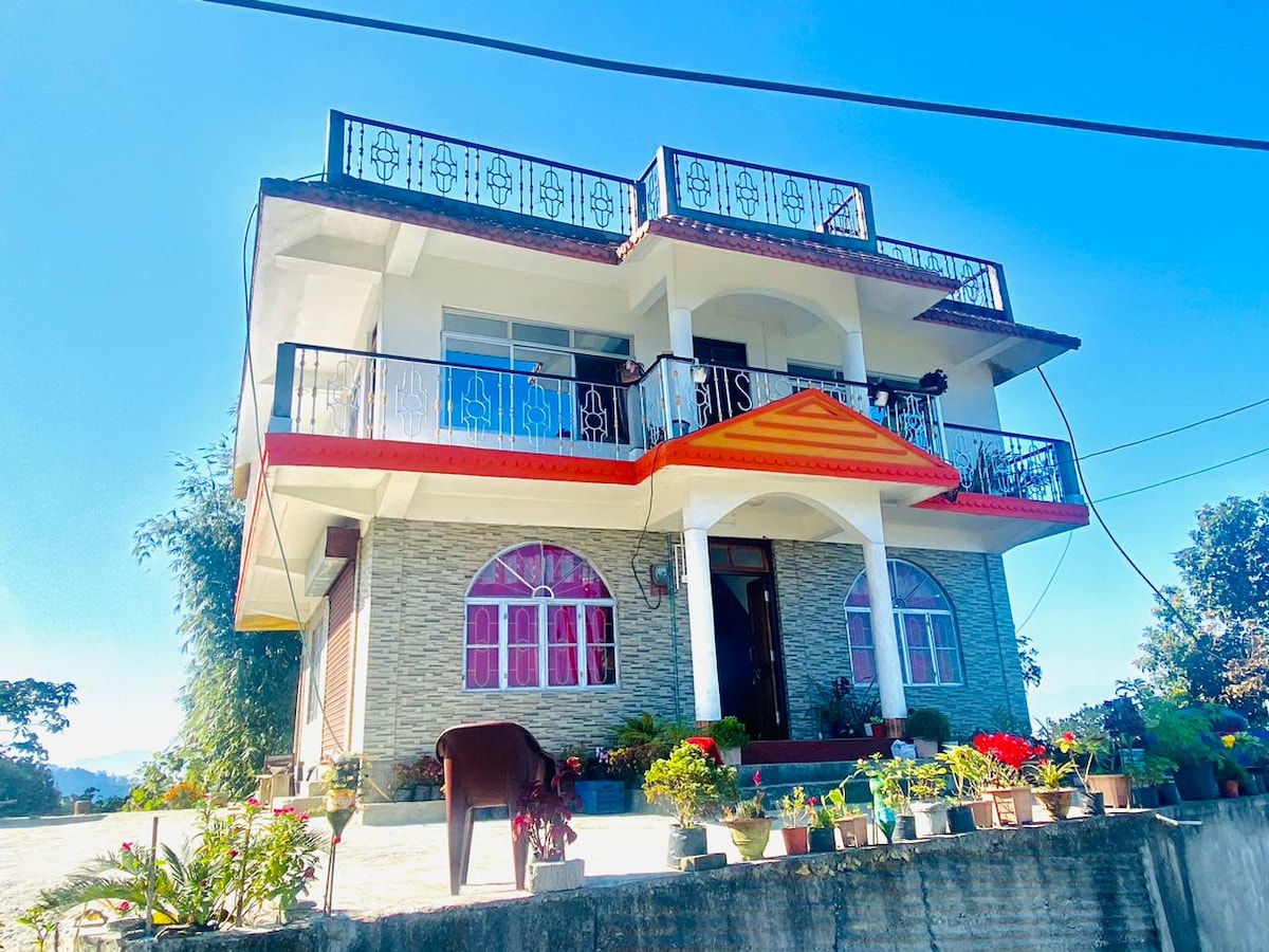 The exterior of a three-story house is visible, featuring a distinct orange accent above the entrance. Flower pots line the ground, adding greenery to the space. Multiple large windows with pink tint reflect sunlight, and a balcony offers ample outdoor seating options.