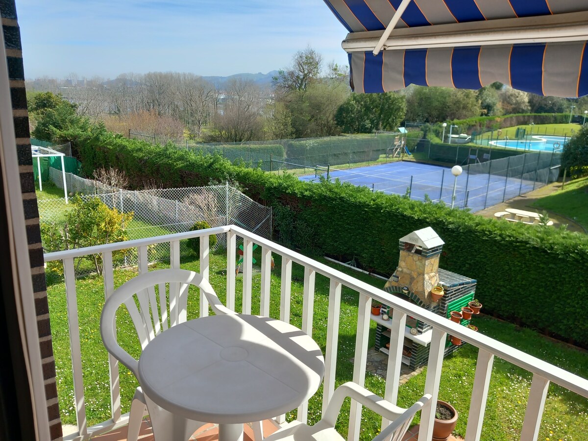 A private balcony is shown, featuring a round table and two white chairs. In the background, neatly maintained tennis courts are visible, surrounded by greenery and fencing. The landscape is illuminated by natural light, creating a serene outdoor space.