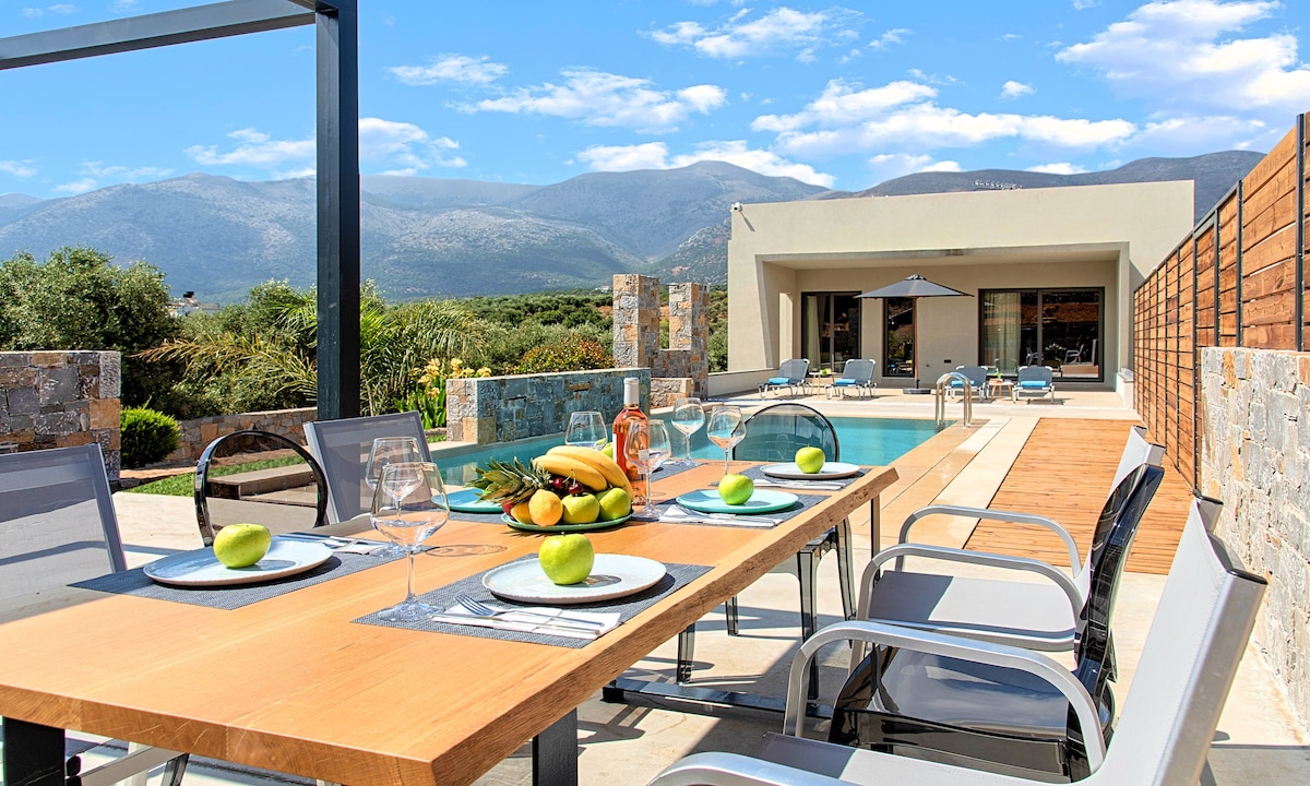 An outdoor dining area features a long wooden table set with plates of fresh fruits and glasses. Lounge chairs surround a pristine pool, with mountains visible in the background under a clear blue sky.