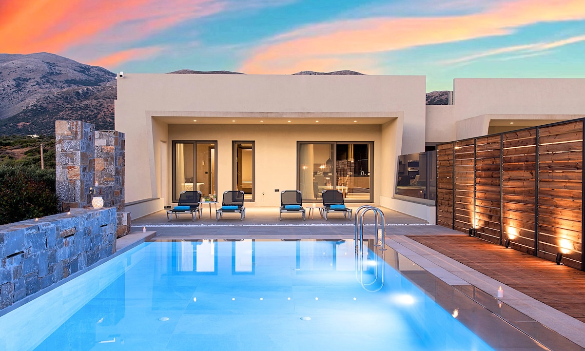 A modern villa is showcased with an inviting swimming pool reflecting the colorful sky at dusk. Sun loungers are arranged beside the pool, and large glass doors open to a spacious indoor area. Natural stone elements are present in the surrounding landscaping.