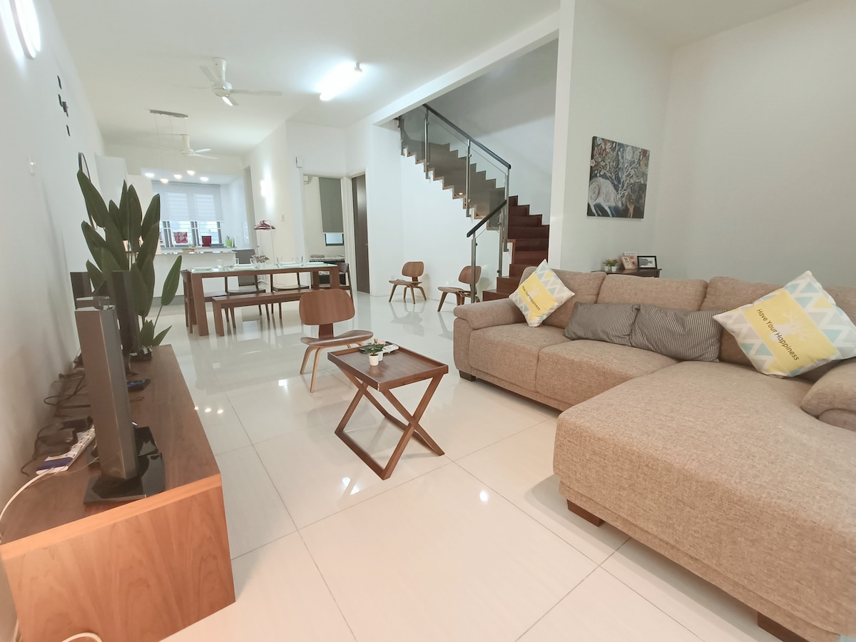 The living area features a spacious sectional sofa accompanied by a coffee table and decorative plants. A dining area with wooden chairs is visible in the background. Bright light fills the room through large windows, complementing the modern design elements and neutral color palette.