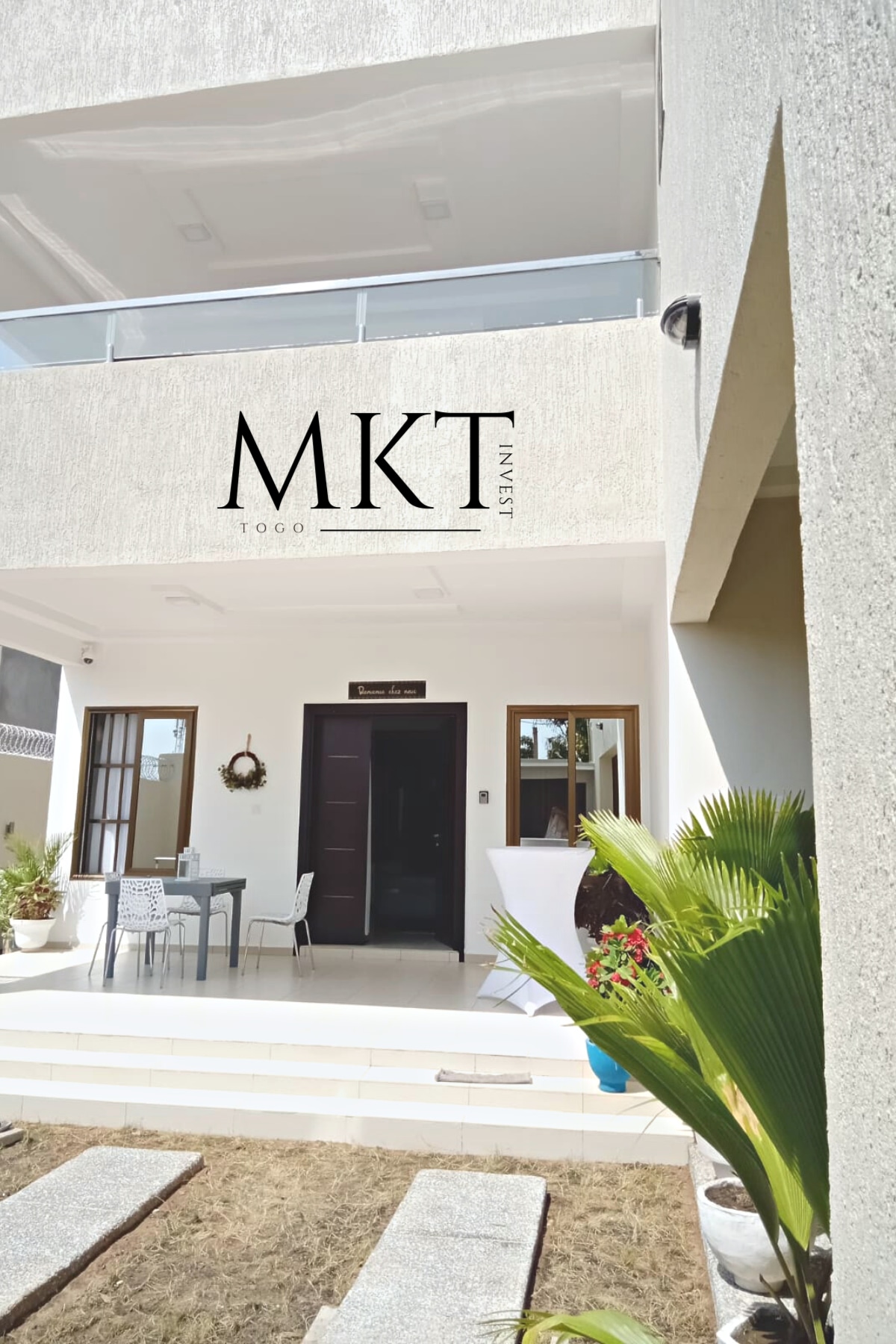 The entrance of the villa is presented, featuring a modern facade with the 'MKT' insignia prominently displayed. A small seating area with a table and chairs is situated on the patio, accompanied by lush greenery and decorative pots, enhancing the approachable vibe.