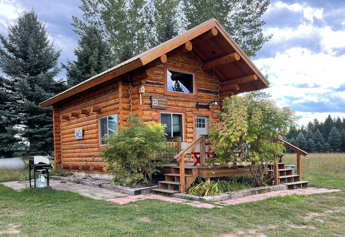 The rustic log cabin is surrounded by tall pines and open fields. A set of stairs leads to a welcoming entrance, while a small porch is adorned with greenery. The structure features large windows, inviting natural light and views of the surrounding landscape.