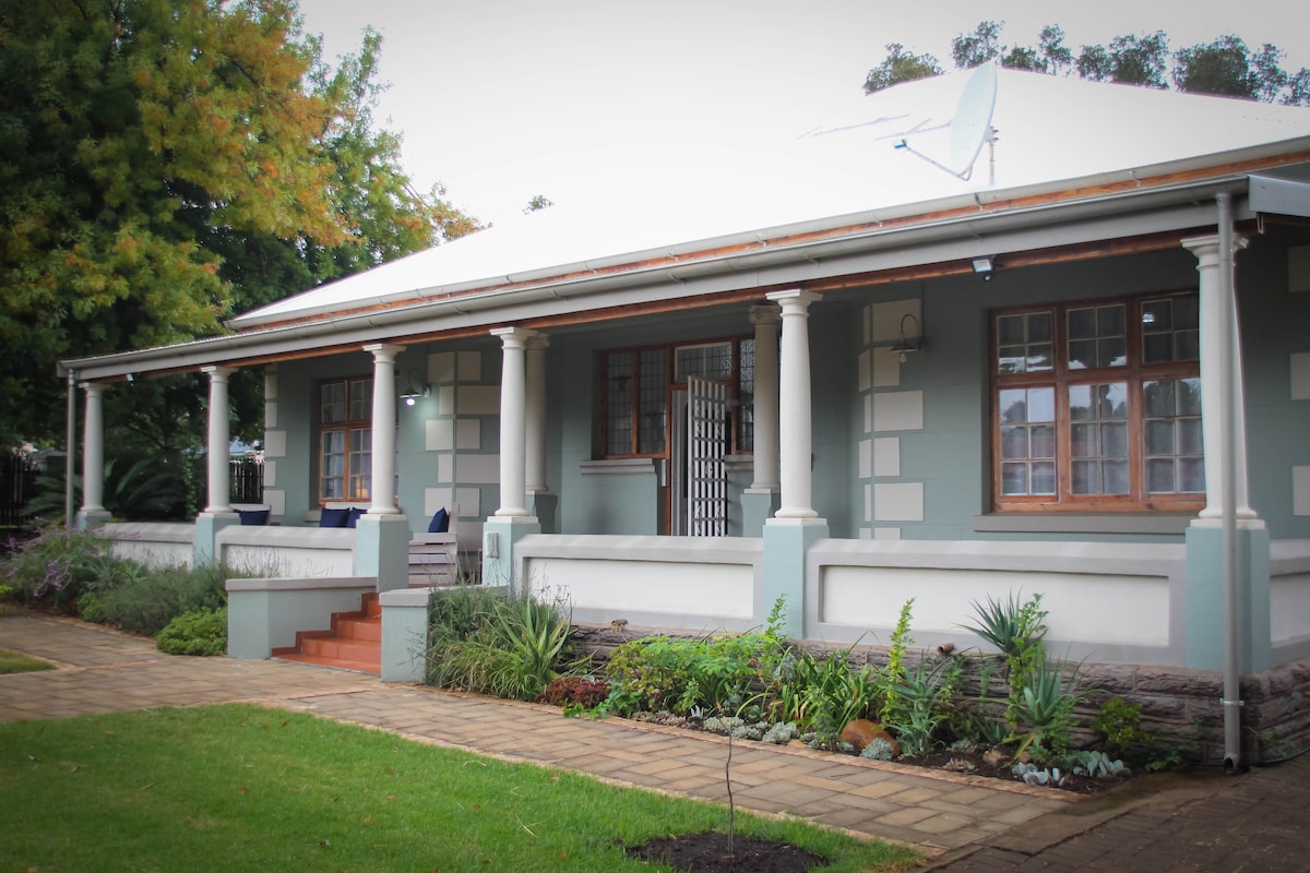 The charming exterior of a spacious home is framed by green gardens and a well-kept lawn. A covered front verandah with white columns provides a welcoming entrance, while steps lead up to the door. A satellite dish is mounted on the roof.
