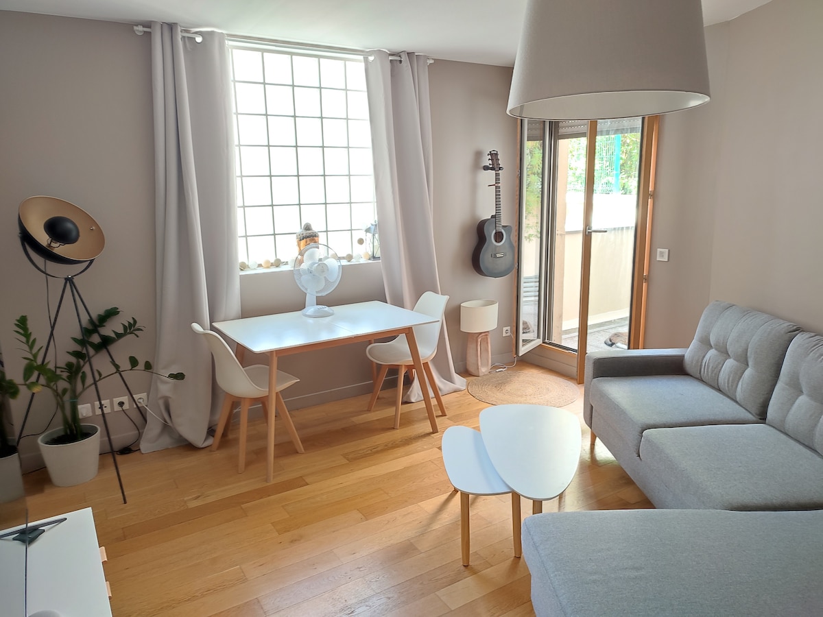A light-filled living area is showcased with a gray sectional sofa and a minimalist coffee table. A dining table with seating for four sits near the window. A fan and a guitar add character to the space, which features warm wood flooring and soft, neutral walls.