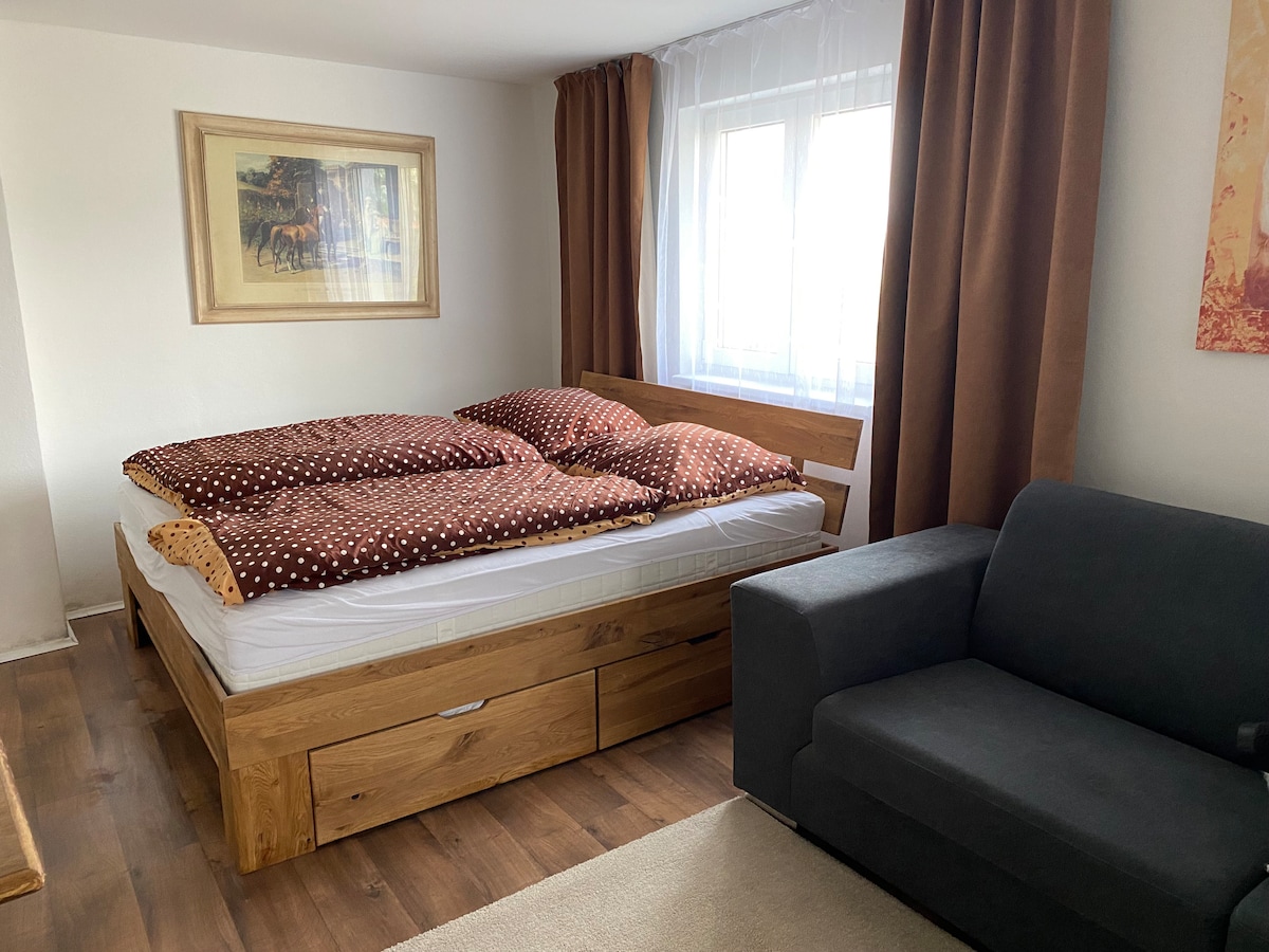 A cozy sleeping area features a spacious bed with a polka-dotted duvet cover. A framed art piece hangs above, and a large window is dressed in light-colored curtains. A comfortable gray sofa is placed nearby, inviting relaxation.