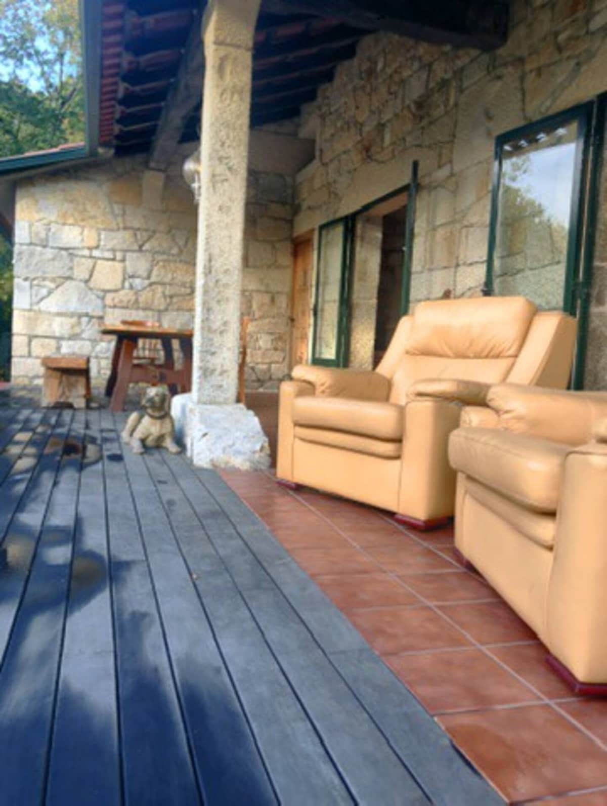 An outdoor area features two warm-toned, cozy armchairs set against a rustic stone wall. A small wooden table is visible nearby, and the flooring alternates between tile and wooden planks. Natural light enters through the large windows, enhancing the inviting space.