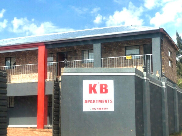 The exterior of a multi-story building is presented, featuring a mix of brick and painted walls. A balcony with railing extends from the second floor, and a prominent sign displays the name 'K'B Apartments' in bold letters, accompanied by contact information.