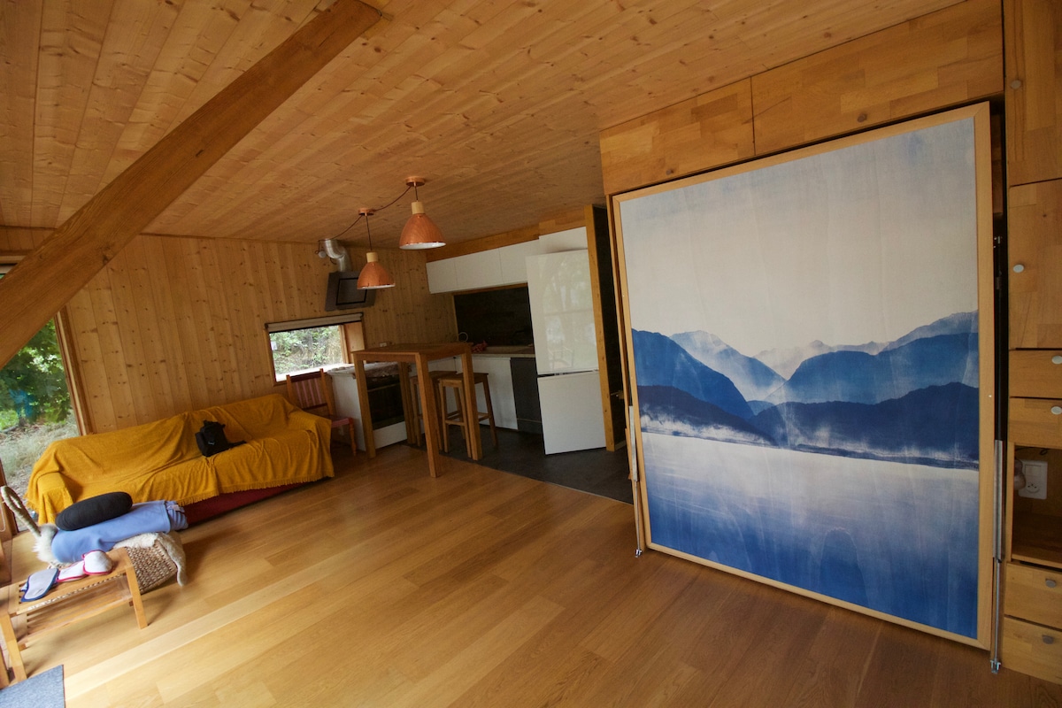 An open living area features wooden walls and a light wooden floor. A yellow sofa is positioned beside a reclining chair. A folding screen with a mountain landscape design separates the space. Hanging copper lights add a touch of warmth above the kitchen area.