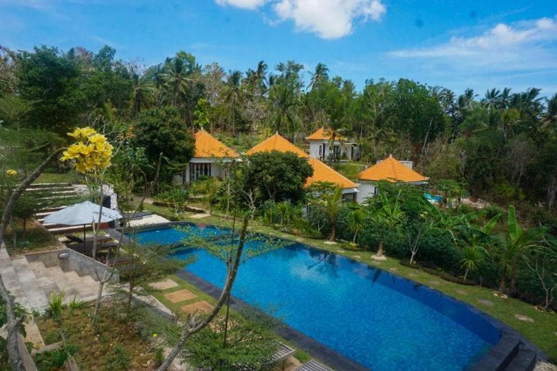 The image features a vibrant pool surrounded by lush greenery and several palm trees. Traditional Balinese villas with orange roofs are visible in the background, set against a clear blue sky. Pathways and garden spaces frame the pool area, inviting outdoor relaxation.