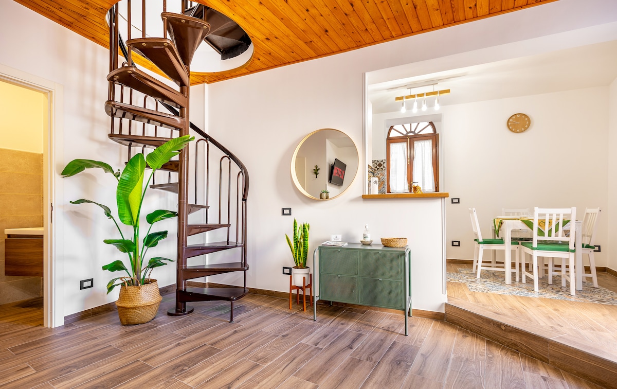 A spiral staircase leads to an upper level, surrounded by natural light filtering through a circular mirror. Potted plants add a touch of greenery, while a wooden sideboard complements the warm tones of the ceiling. A dining area can be glimpsed in the background.