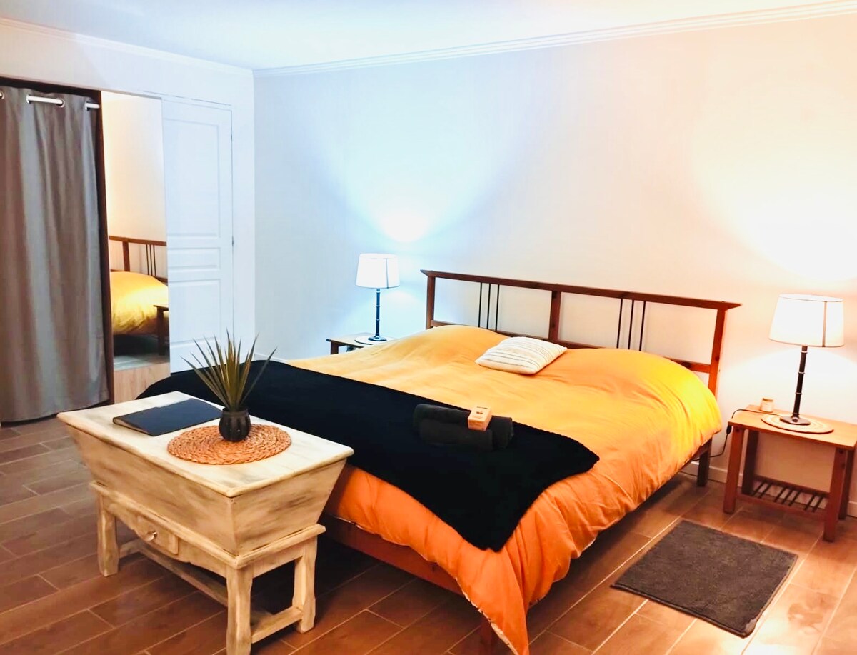 A spacious bedroom features a large orange comforter covering a bed with a wooden headboard. Two bedside lamps provide gentle lighting, while a small wooden table holds a plant and items. A dark rug sits on the tiled floor, and a doorway is visible leading to another room.