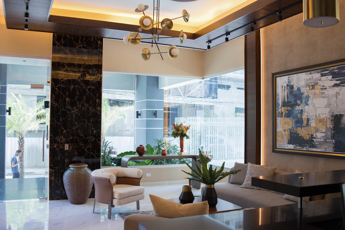 The welcoming lobby features a modern design with sleek furnishings. A contemporary chandelier hangs from the ceiling, while a large artwork adorns the wall. Greenery is visible outside through expansive glass doors, and seating options are provided with plush chairs and a sectional sofa.