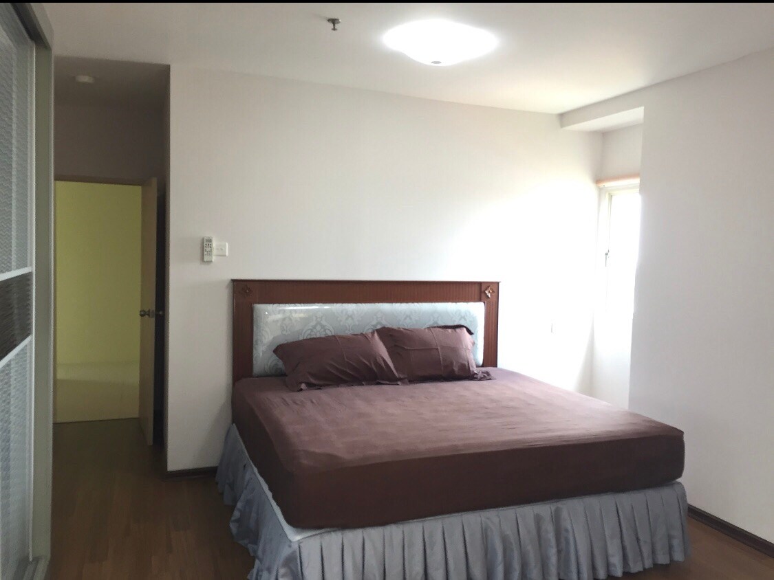 A spacious sleeping area features a large, unmade bed with a muted brown coverlet and matching pillows. Natural light enters through a nearby window, illuminating the room's simple design and wooden floors. A door on the left leads to another part of the apartment.