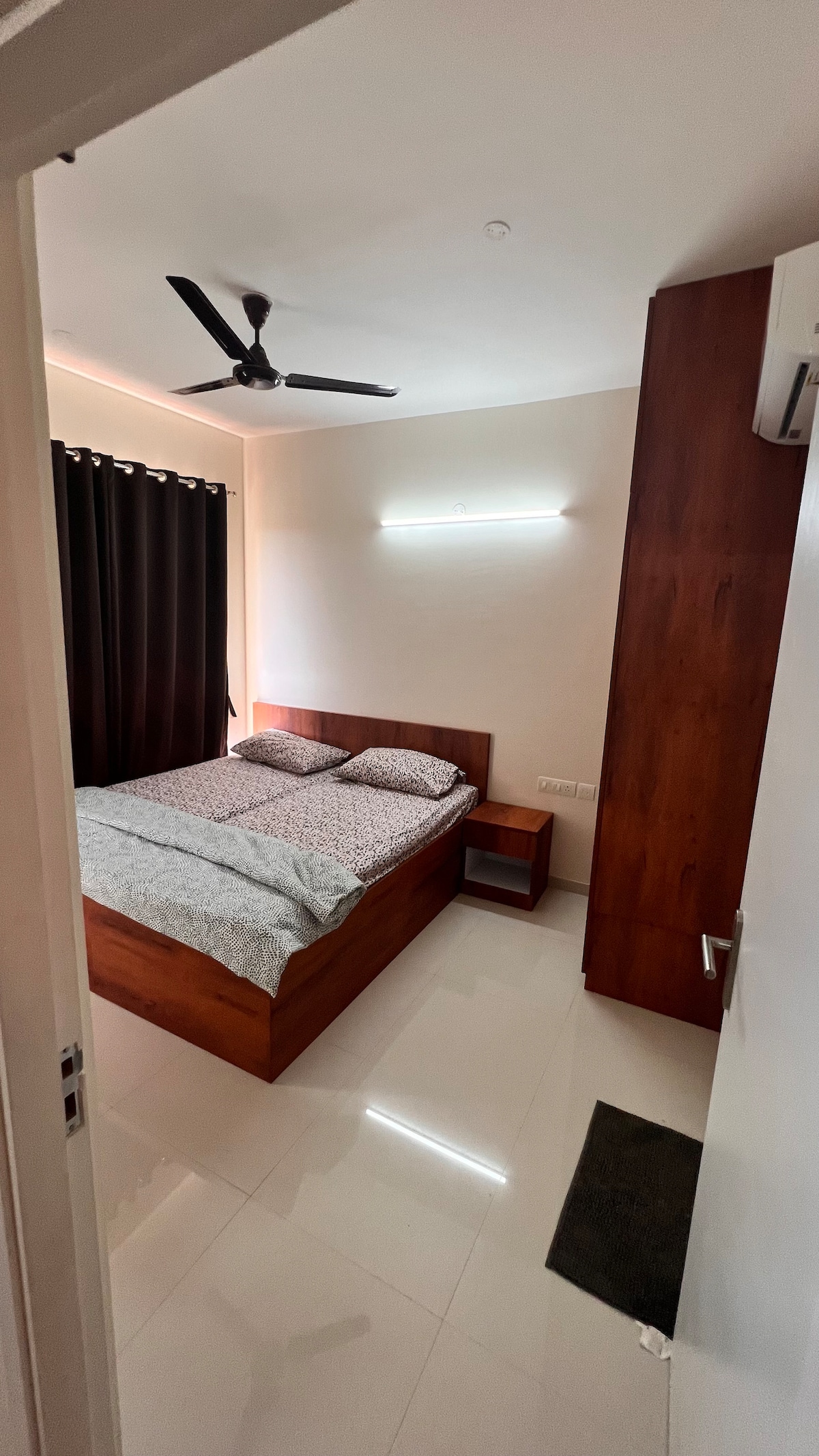 A well-lit bedroom features a wooden bed with a patterned grey and white duvet. A small bedside table complements the bed, and a large closet stands nearby. Dark curtains help to control light, and a ceiling fan contributes to the room’s comfort.