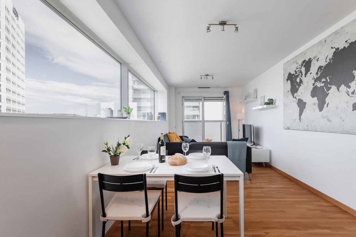 A bright dining area is visible, featuring a white table set for two with glassware and a loaf of bread. A cozy living space is located in the background, with a dark sofa, a television, and a decorative map on the wall under ambient lighting.