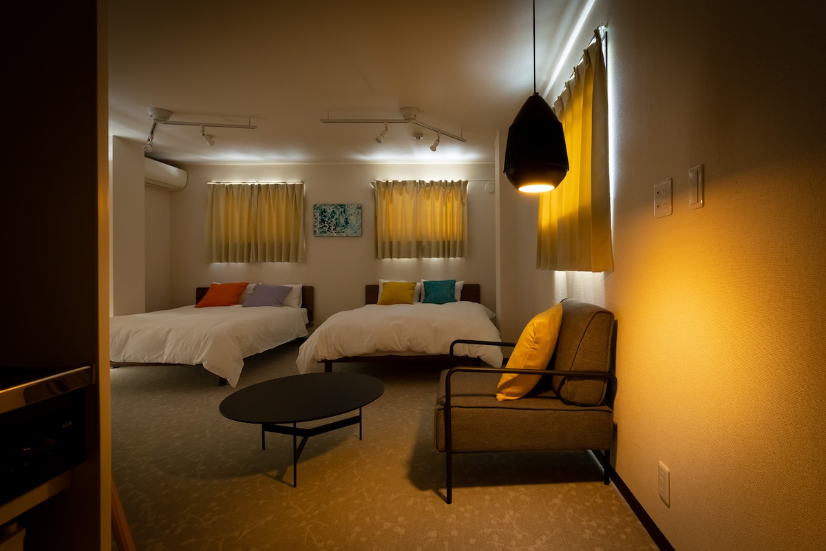 A well-furnished room features two spacious beds with colorful accent pillows. Soft light filters through sheer curtains at the windows. A comfortable chair is positioned near a low coffee table, creating a balanced and inviting seating area.
