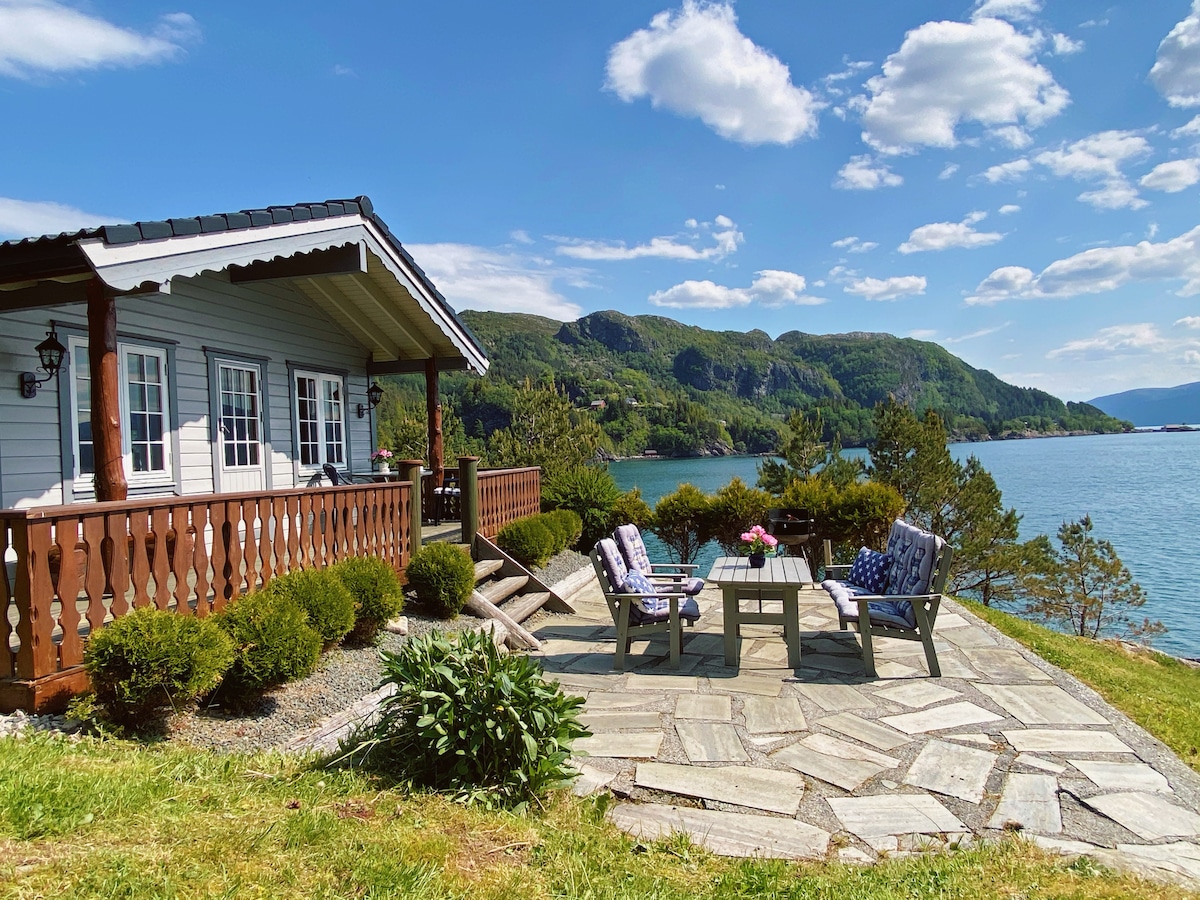 A scenic outdoor terrace is featured in this image, with comfortable seating arrangements and a stone pathway leading to lush greenery. The backdrop showcases a tranquil fjord and rolling hills under a partly cloudy sky.