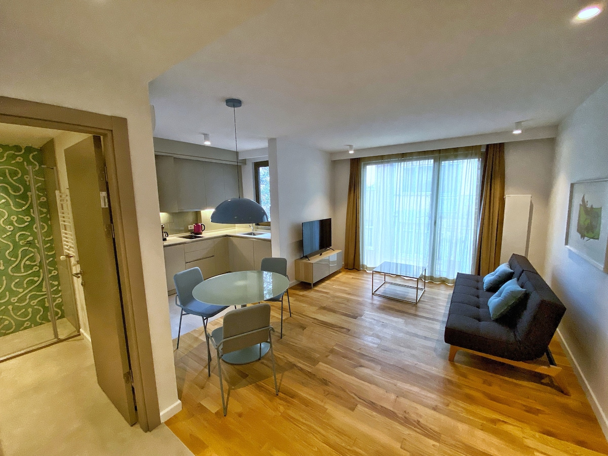 A modern living space is highlighted by a combination of light wood flooring and neutral-colored walls. A circular dining table is set with four chairs. A sofa in front of a wall-mounted television offers comfortable seating. Floor-to-ceiling windows allow natural light to fill the room.