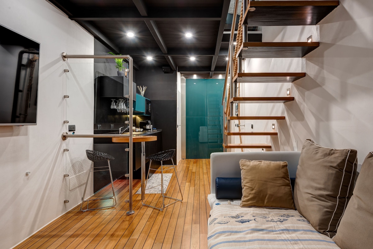The interior features a sleek and modern design, with wooden flooring and a cozy seating area. A compact kitchenette is visible, accompanied by a dining table and stylish bar stools. An open staircase leads to the upper sleeping area, illuminated by soft lighting.
