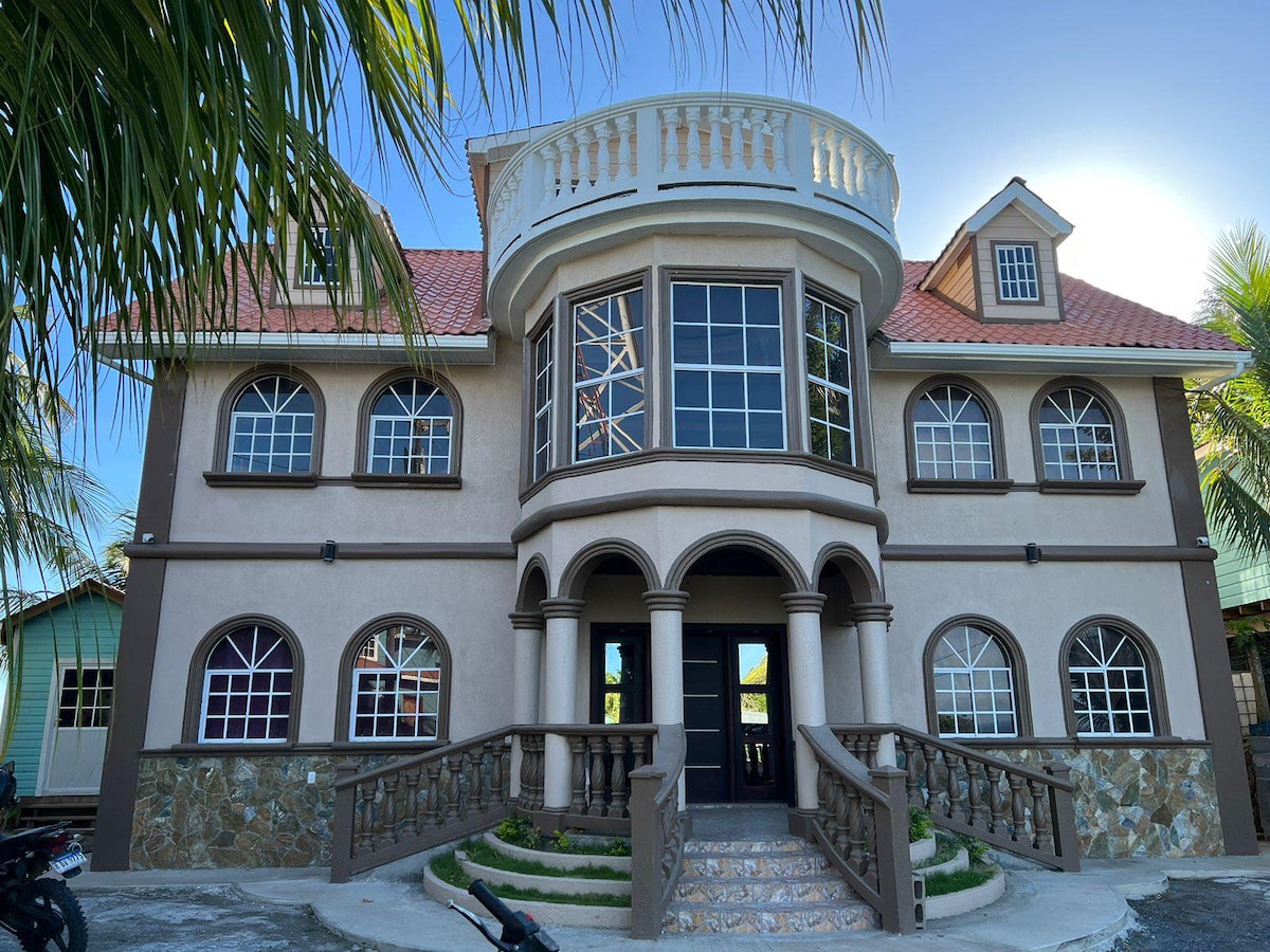 The exterior of a large, multi-story house is presented, featuring a round central structure with a balcony and multiple arched windows. The façade combines light and dark tones, and a staircase with a decorative railing leads to the entrance. Lush greenery is visible in the background.