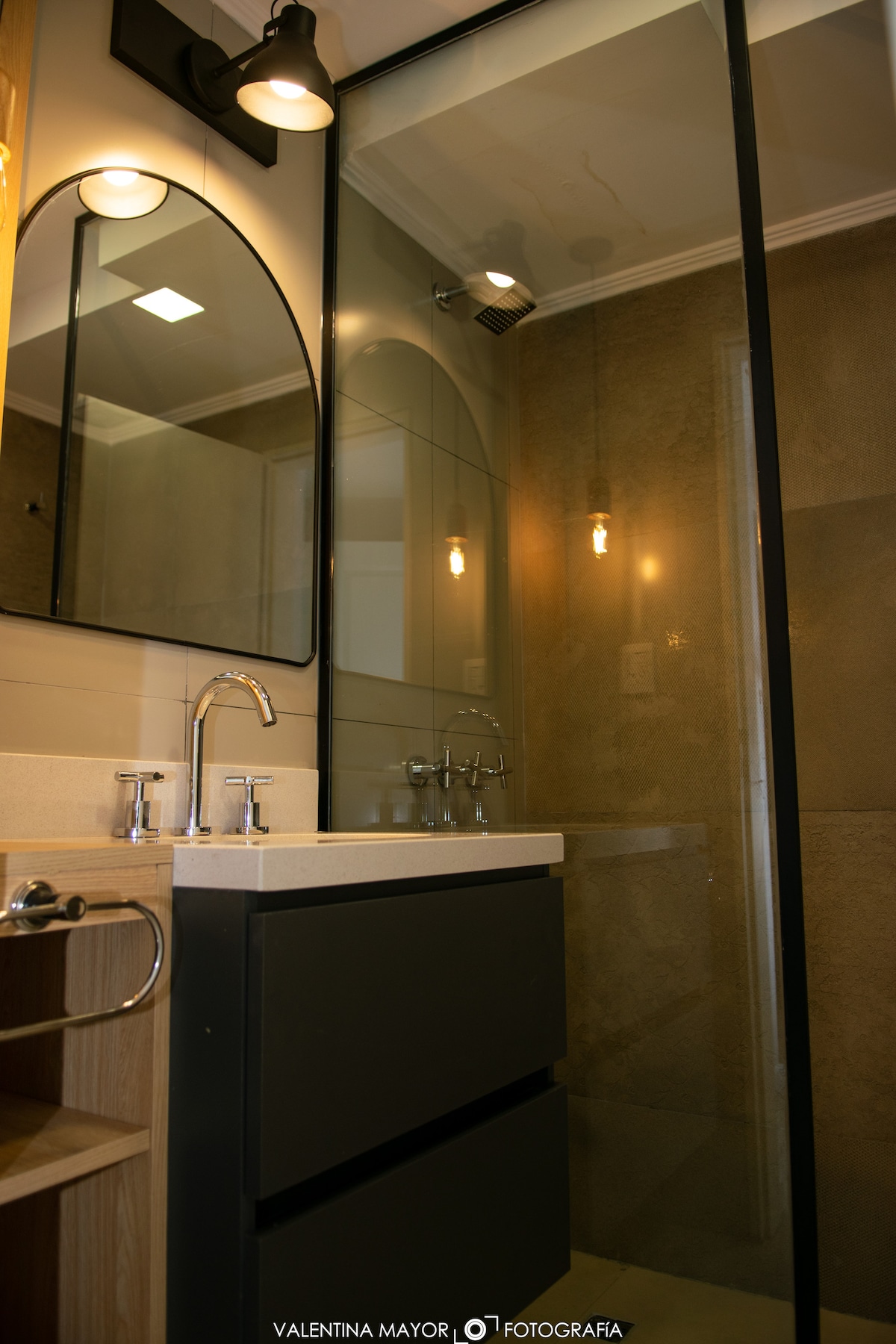 A modern bathroom showcases a sleek, dark cabinetry with a minimalist sink and polished faucets. Two rounded mirrors reflect soft lighting from above, and a glass shower enclosure is visible. Neutral-colored tiles create a clean and functional space.